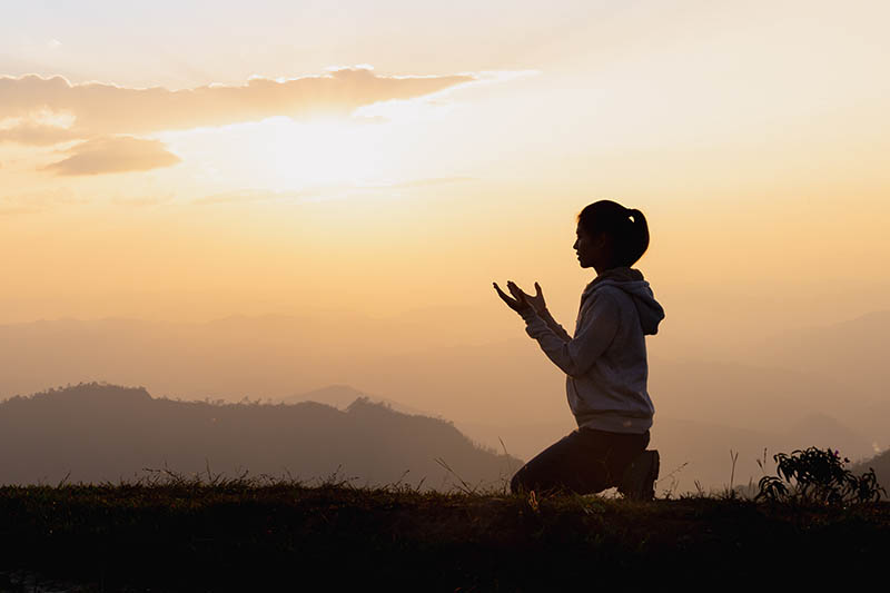 A young woman kneels and prays at sunset while overlooking ridges of hills and valleys