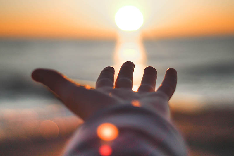Hand reaching out to the sun as it sets over the ocean