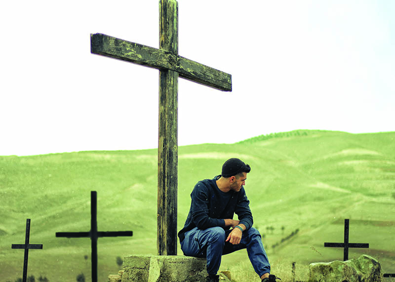 Middle Easten young man sitting among crosses on a green hillside