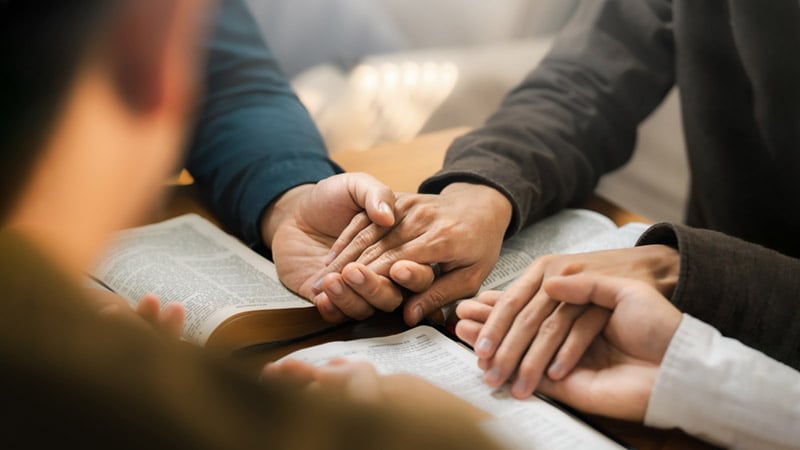A small circle of people with open Bibles, holding hands, and praying. 