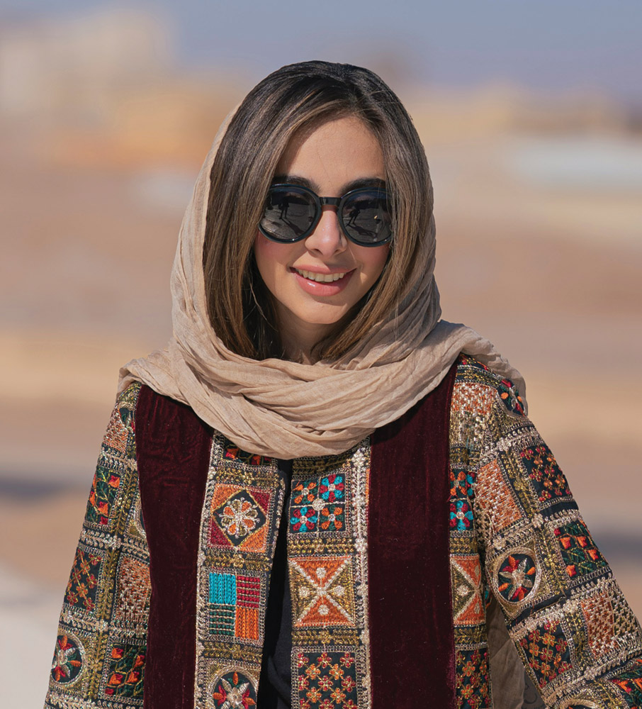 A middle-eastern young woman wearing a colorful jacket and sunglasses smiles
