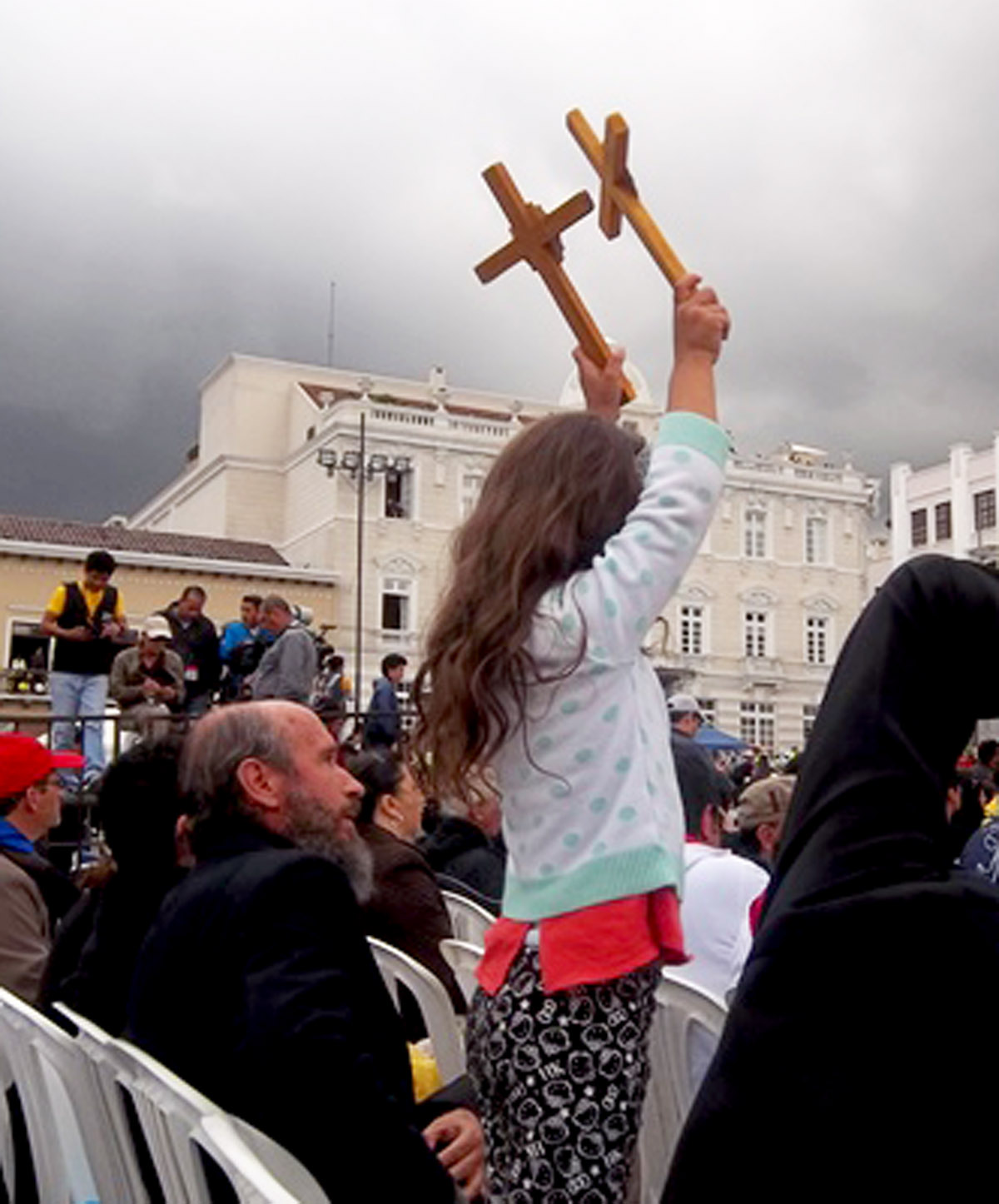A young girl raises a pair of crosses while seeing Pope Francis from a distance. Photo credit: Sondra Holden